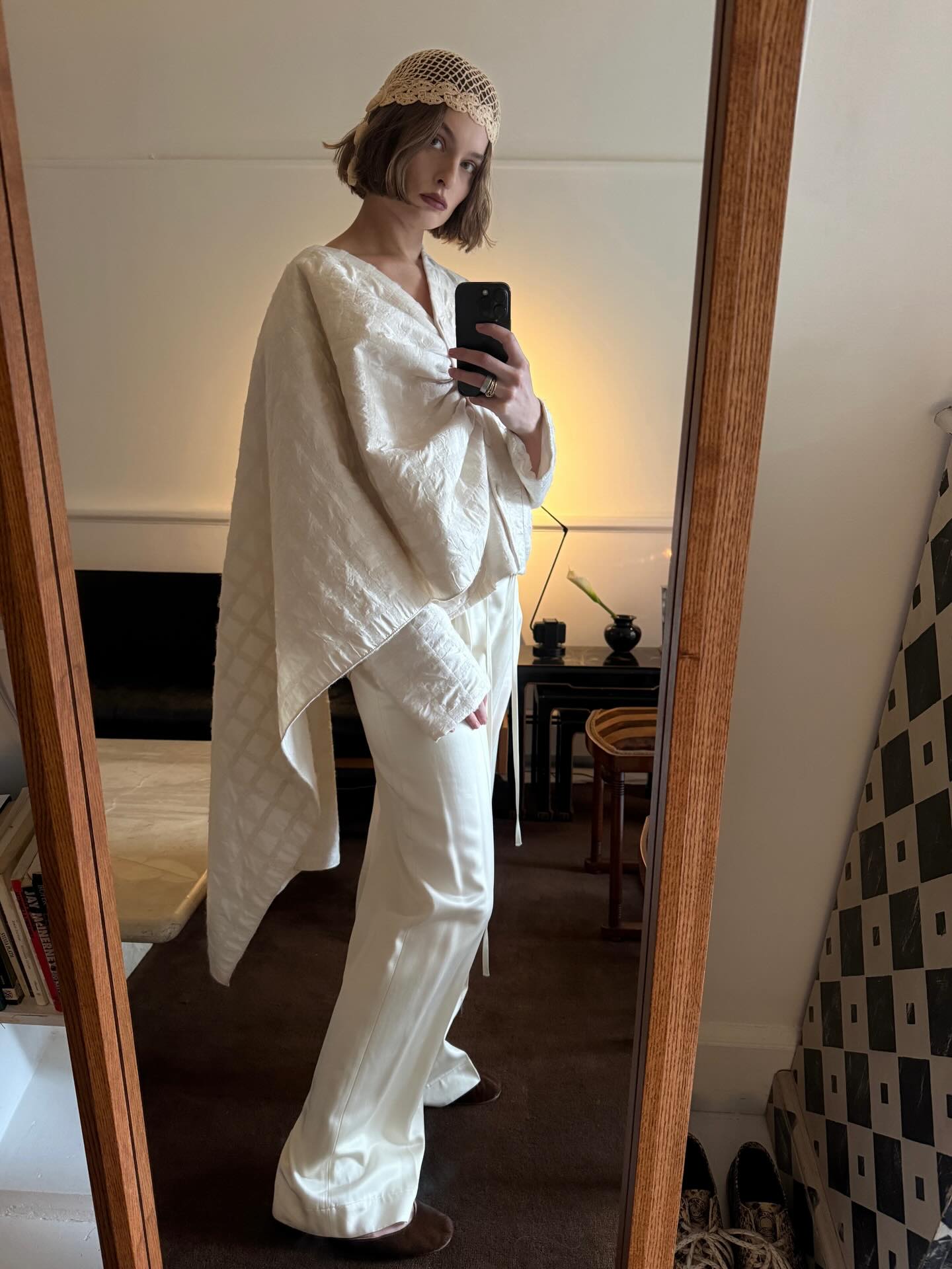 An image of an influencer wearing a white cap blouse, white trousers and a beige crochet skull cap taking a photo in a mirror.