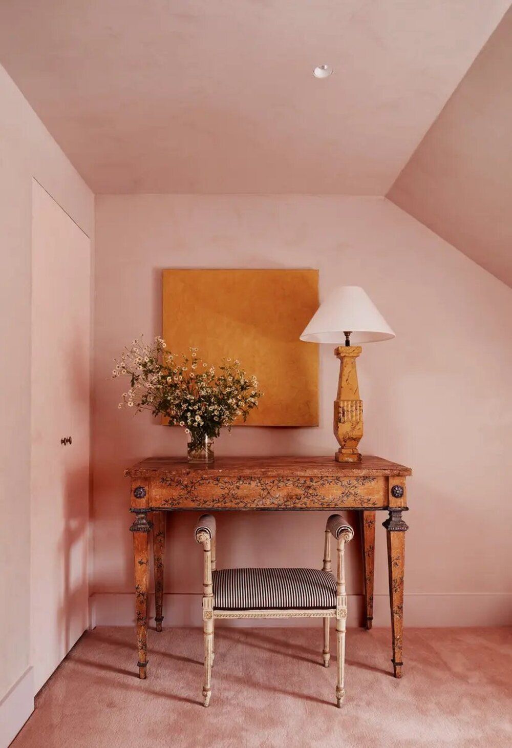 pink color drenched bedroom with plaster pink walls, pink carpet, antique desk, antique stool, vase of flowers, antique desk lamp, and orange artwork