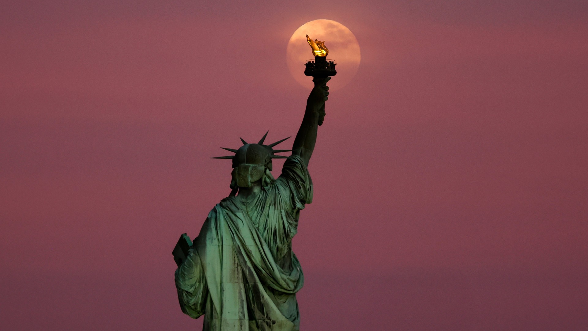 The Statue of Liberty in New York is pictured with the orange full 'Flower Moon' rising behind the statue's raised torch. The scene is cloudy, and red hued.
