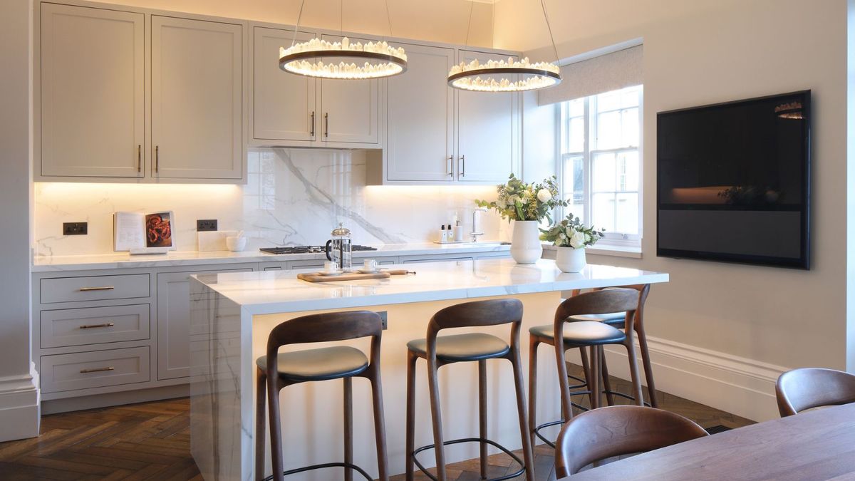 A modern kitchen with a white marble island featuring waterfall edges. Three dark brown barstools with curved backs and black metal footrests are lined up along the island. Above, two circular chandeliers with small crystal-like details hang.