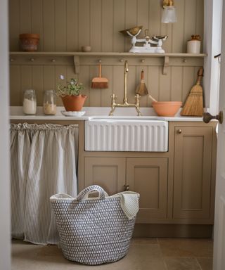 A kitchen with light brown wall paneling and cabinets, white countertops and a sink, a brass tap, and a storage basket