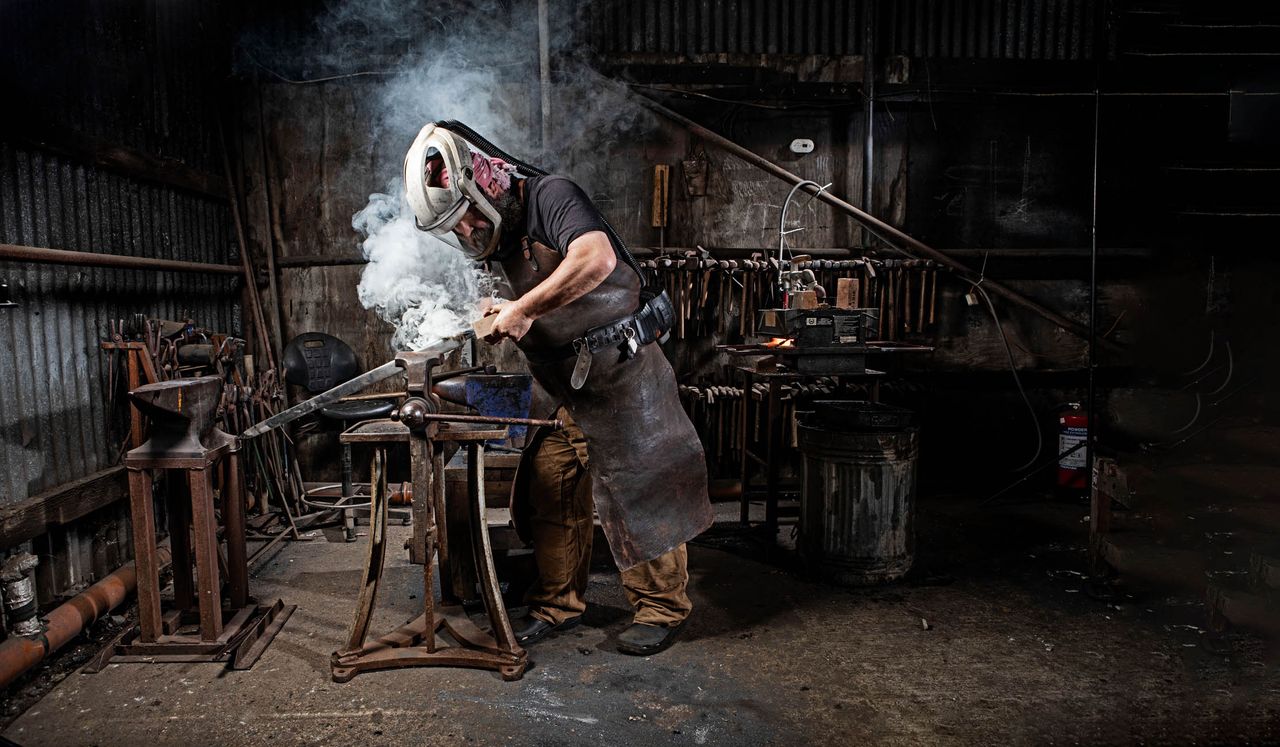 Owen Bush, bladesmith, forging a sword. The smoke billows when he heats up the end of the sword and slides the wooden handle on. The item on his face is called a powered respirator. Photographed at his workshop by Richard Cannon.