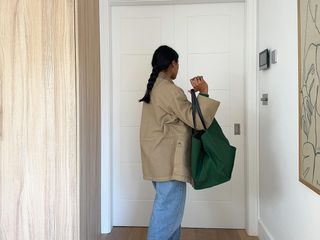 A woman walking toward her door wearing a tan oversize coat and loose, light-wash jeans carrying a green travel bag