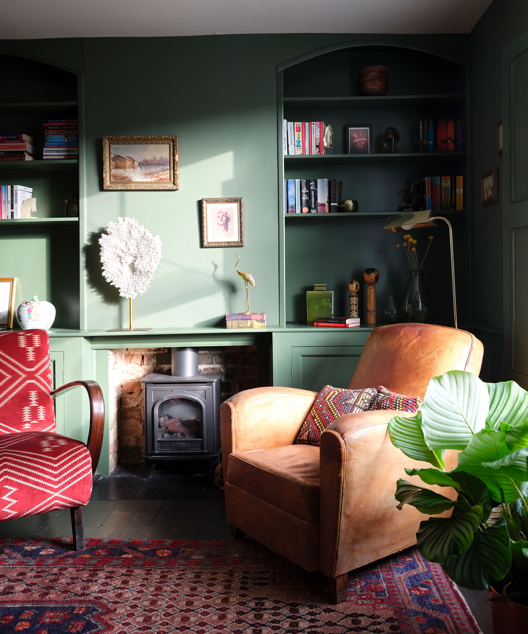 A green living room with built-in alcove storage either side of a vintage fireplace, leather armchair and red and blue patterned Persian rug.