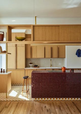 An oxblood tiled kitchen island with a stovetop and kettle. Above the island is a mix of open and closed wooden shelving, and opposite the island are oak cabinets.