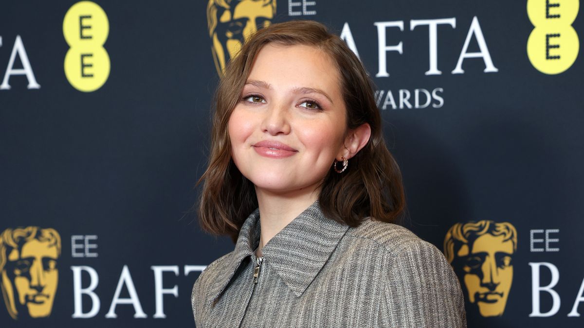 Mia McKenna-Bruce standing in front of a BAFTA branding board and smiling while wearing a smart grey dress with a zip up front and a big collar 