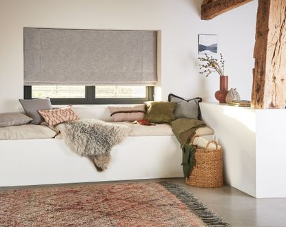 A cosy living room with a corner sofa dressed with throws and cushions and a window with grey blinds