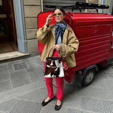 Fashion influencer Vicky Montanari wearing Maison Margiela Tabi ballerina flats, red jeans, a cropped trenchcoat, blue scarf, sunglasses, and carrying a Tory Burch cow print bag.