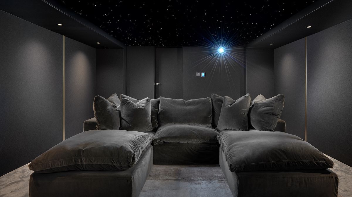 A photograph of a luxury, custom-installed home cinema with a huge projector screen, lush u-shaped sofa, grey walls and lamps