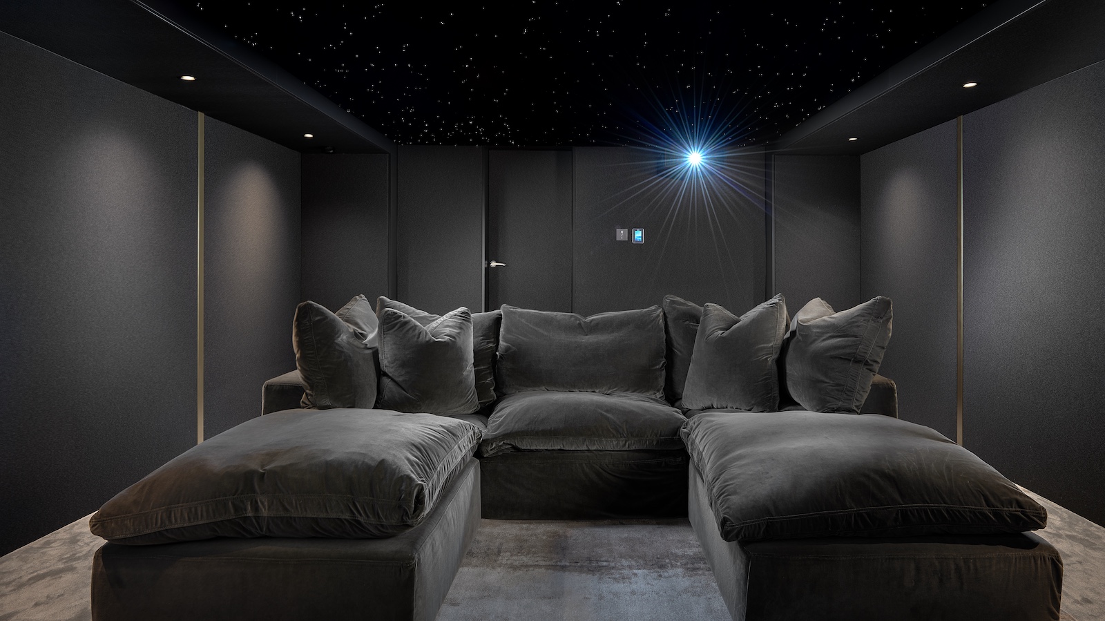 A photograph of a luxury, custom-installed home cinema with a huge projector screen, lush u-shaped sofa, grey walls and lamps