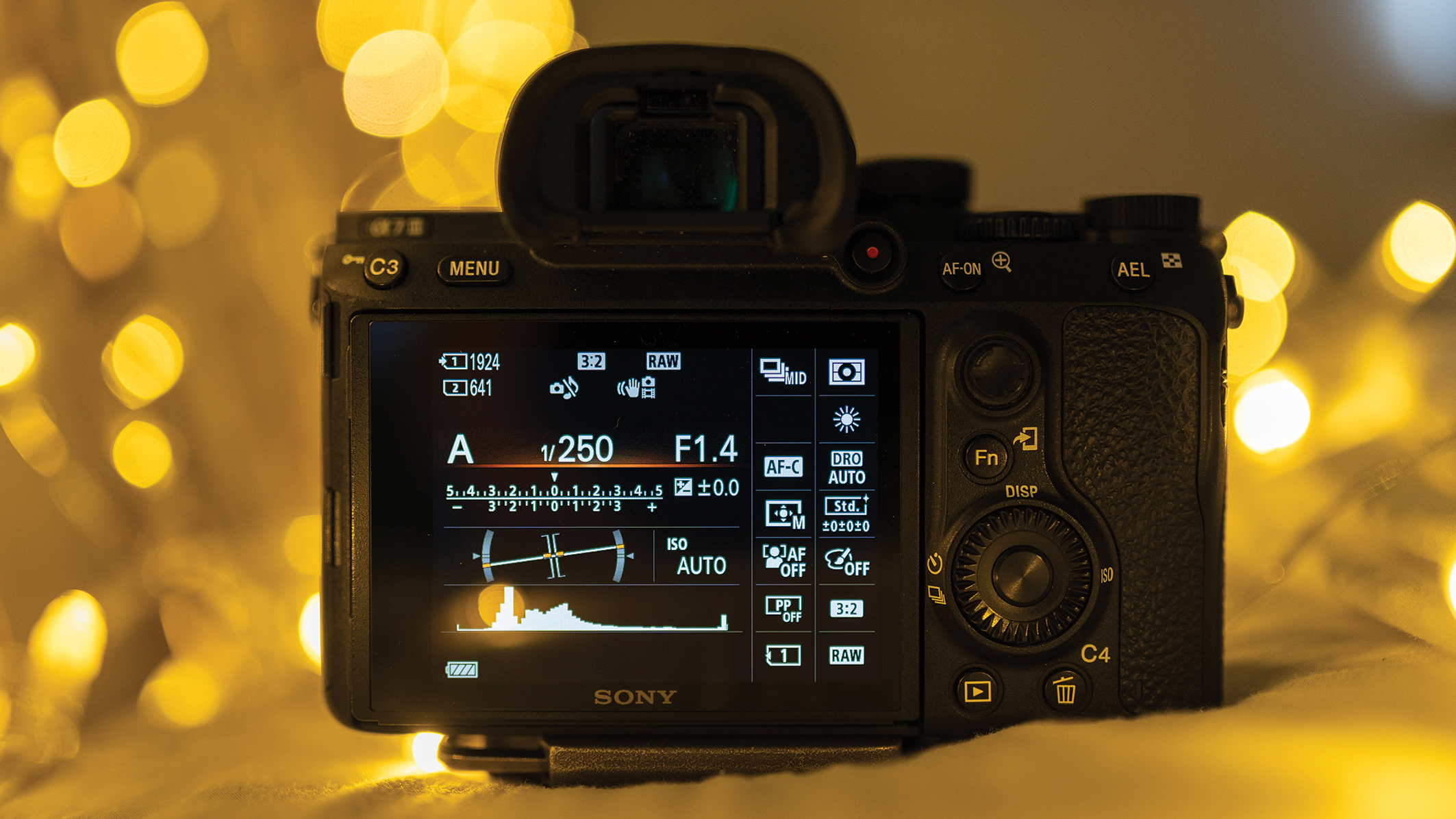 Close-up of a digital camera's back, displaying settings like aperture and ISO on its screen. Warm, blurred bokeh lights create a cozy ambiance