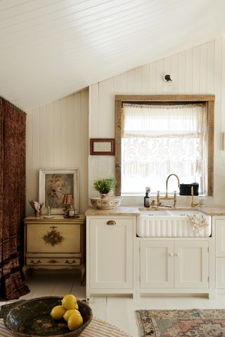A white kitchen with fluted sink, brass taps, off-white shaker cabinets and shiplap walls, with wooden frame to large window