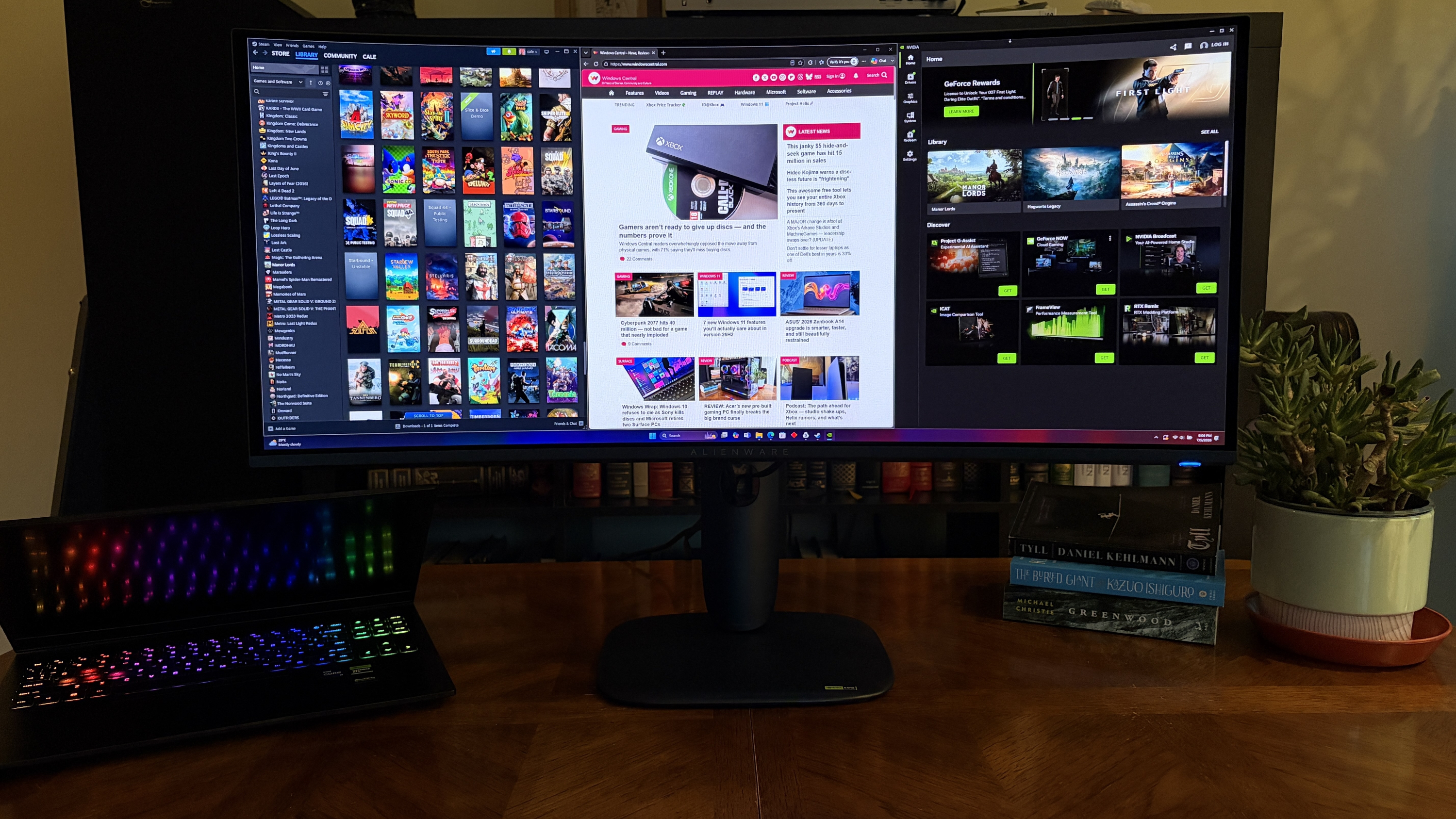 A workspace with an Alienware AW3426DW monitor displaying gaming content, a black laptop with a rainbow keyboard, and a small green plant next to stacked books on a wooden table.
