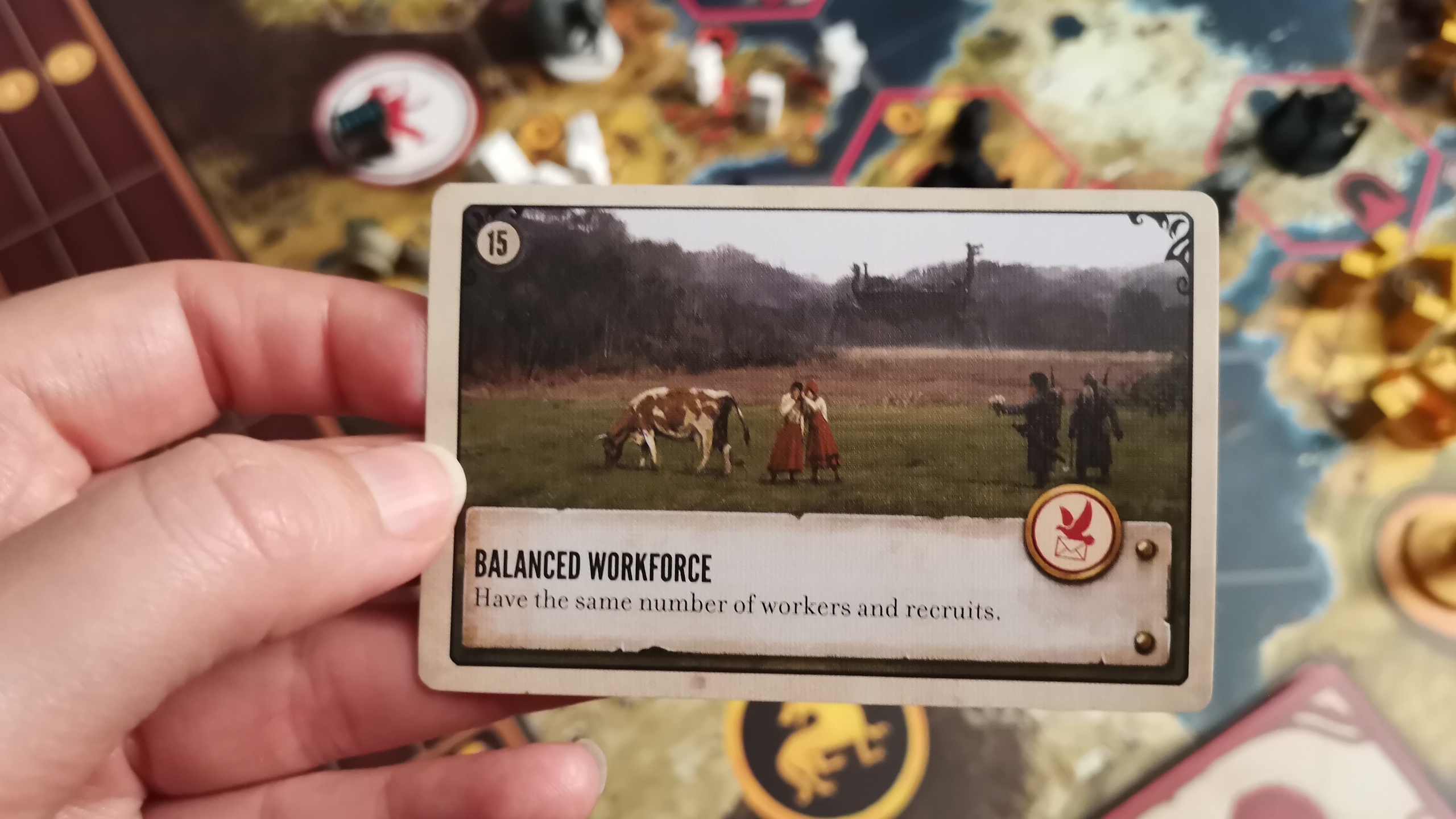 A hand holds a 'balanced workforce' Scythe card, with the board in the background