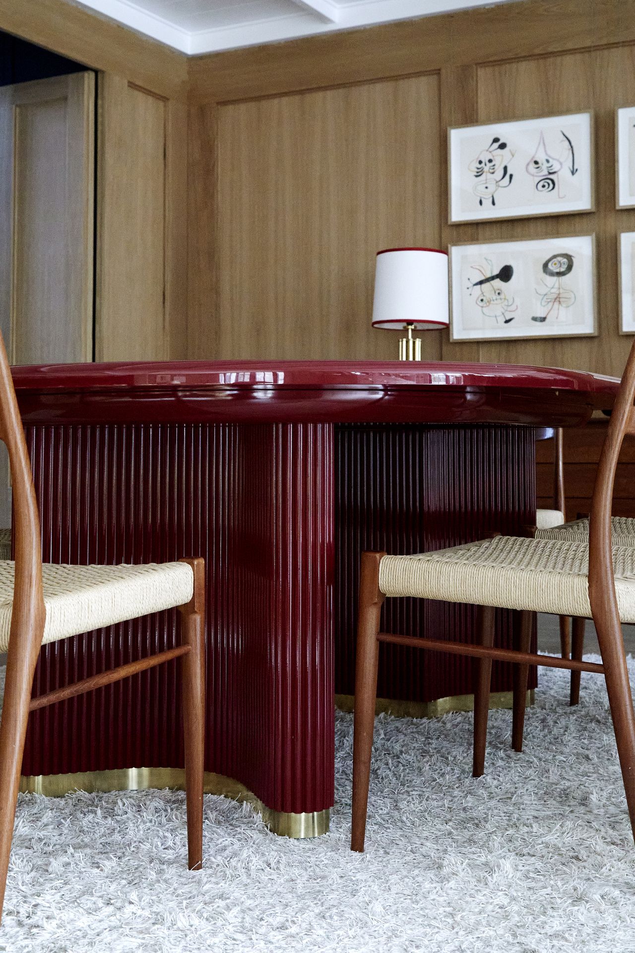 Close-up image that is eye-level with a glossy, red dining table. There is a fluffy white rug on the floor and two wooden dining chairs in view.