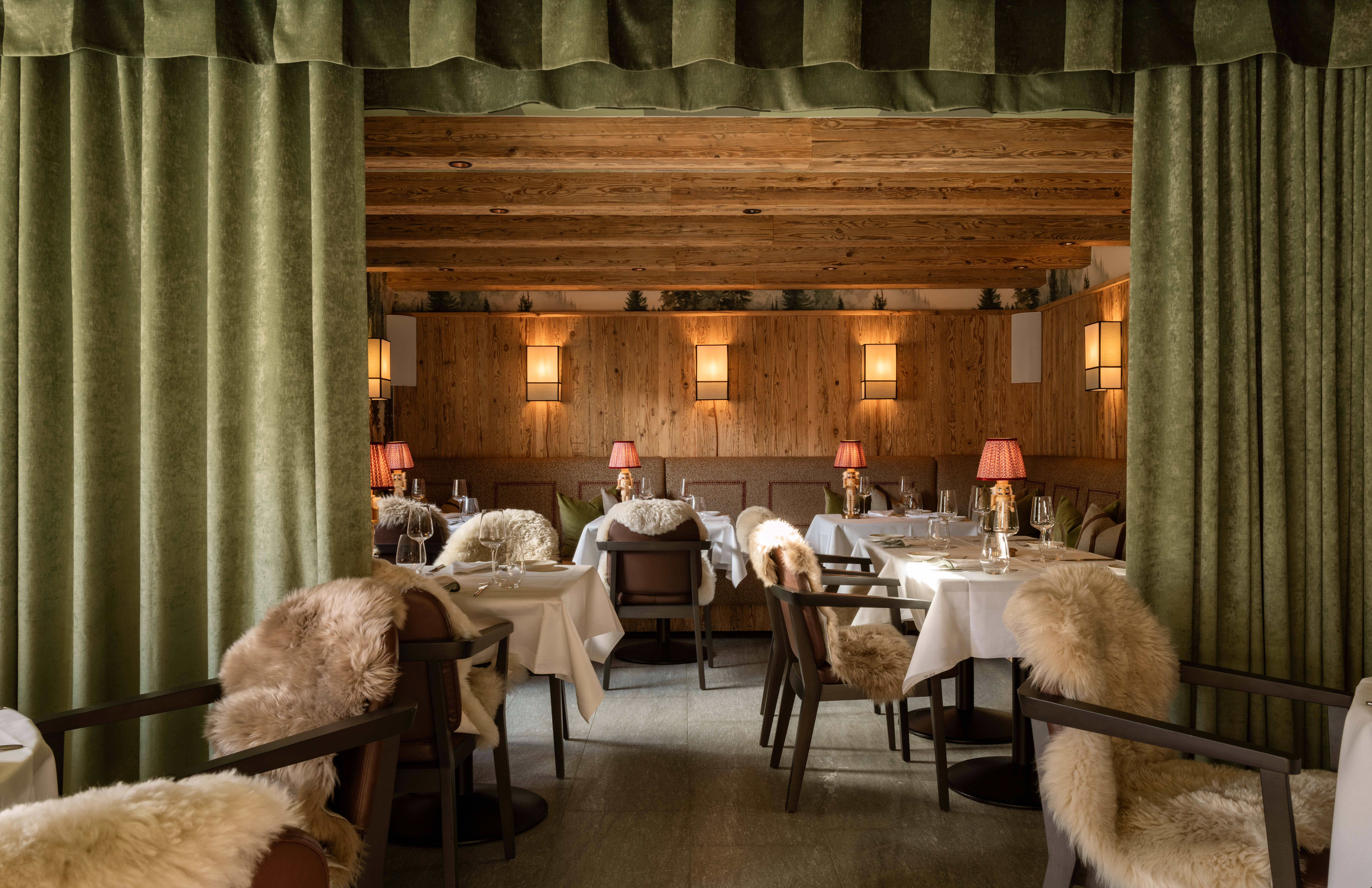 A luxurious alpine retreat's restaurant is decorated with pale wood-paneled walls, dark wood chairs covered in sheepskin blankets, Art Nouveau table lamps, golden lighting, and green curtains.