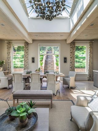 Open plan living and dining room with natural light with a roof lantern, wooden floor, rug, sofas and chairs and chandelier, and a view to the garden.