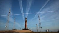 A large orange and white rocket sits on a launch pad with streaks of white clouds behind it