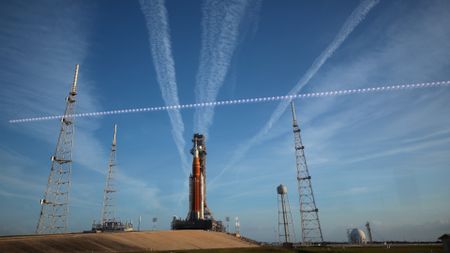 A large orange and white rocket sits on a launch pad with streaks of white clouds behind it