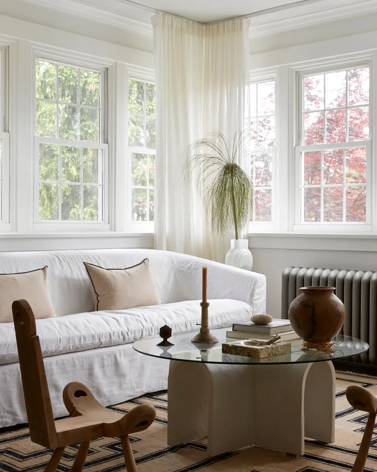 White living room with white sofa, white and glass topped sculptural coffee table, wooden armchairs and black and brown geometric area rug