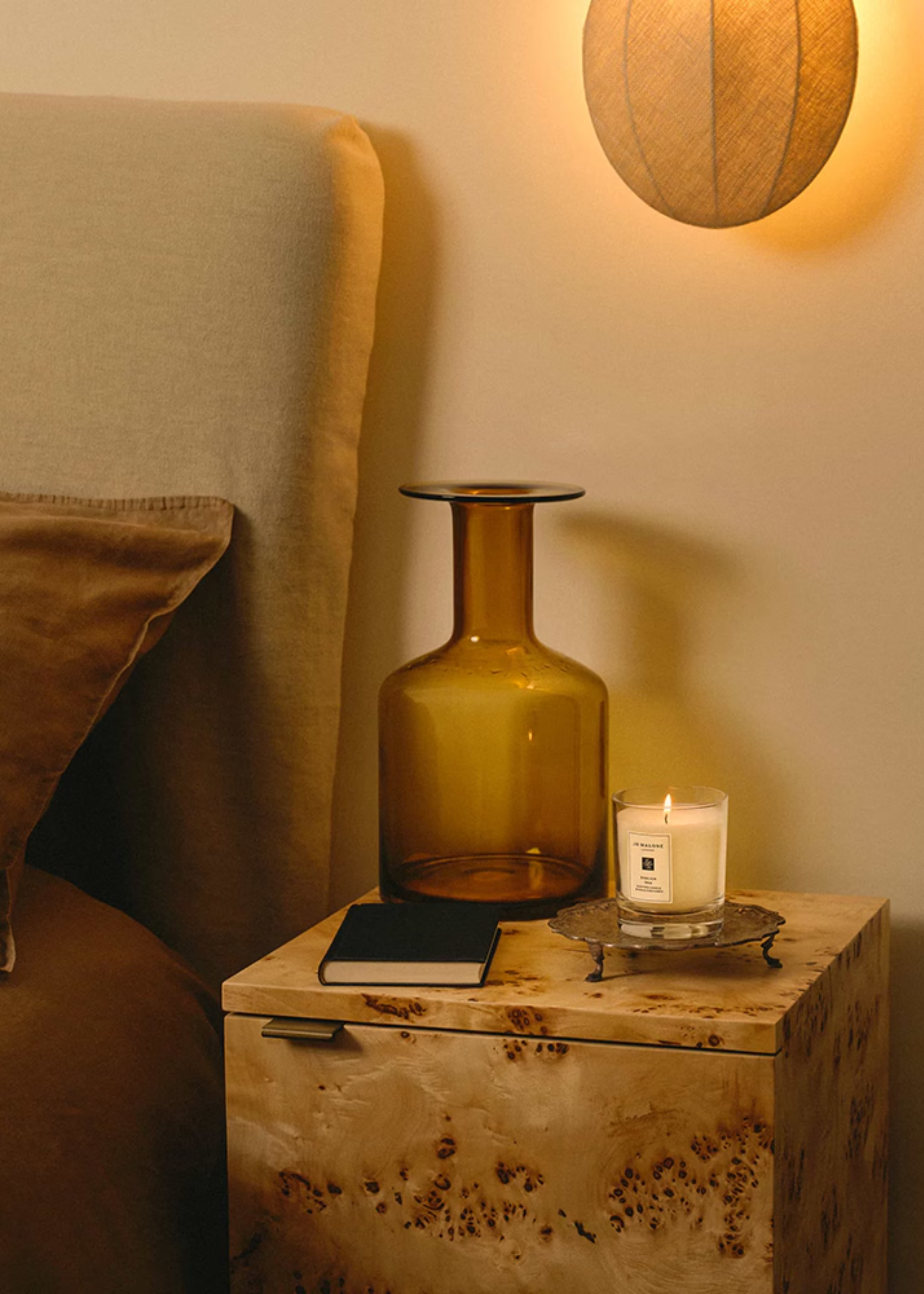 A bedroom with a burl wood side table featuring a large vase, a book, a Jo Malone candle on a metal stand and a woven sconce above