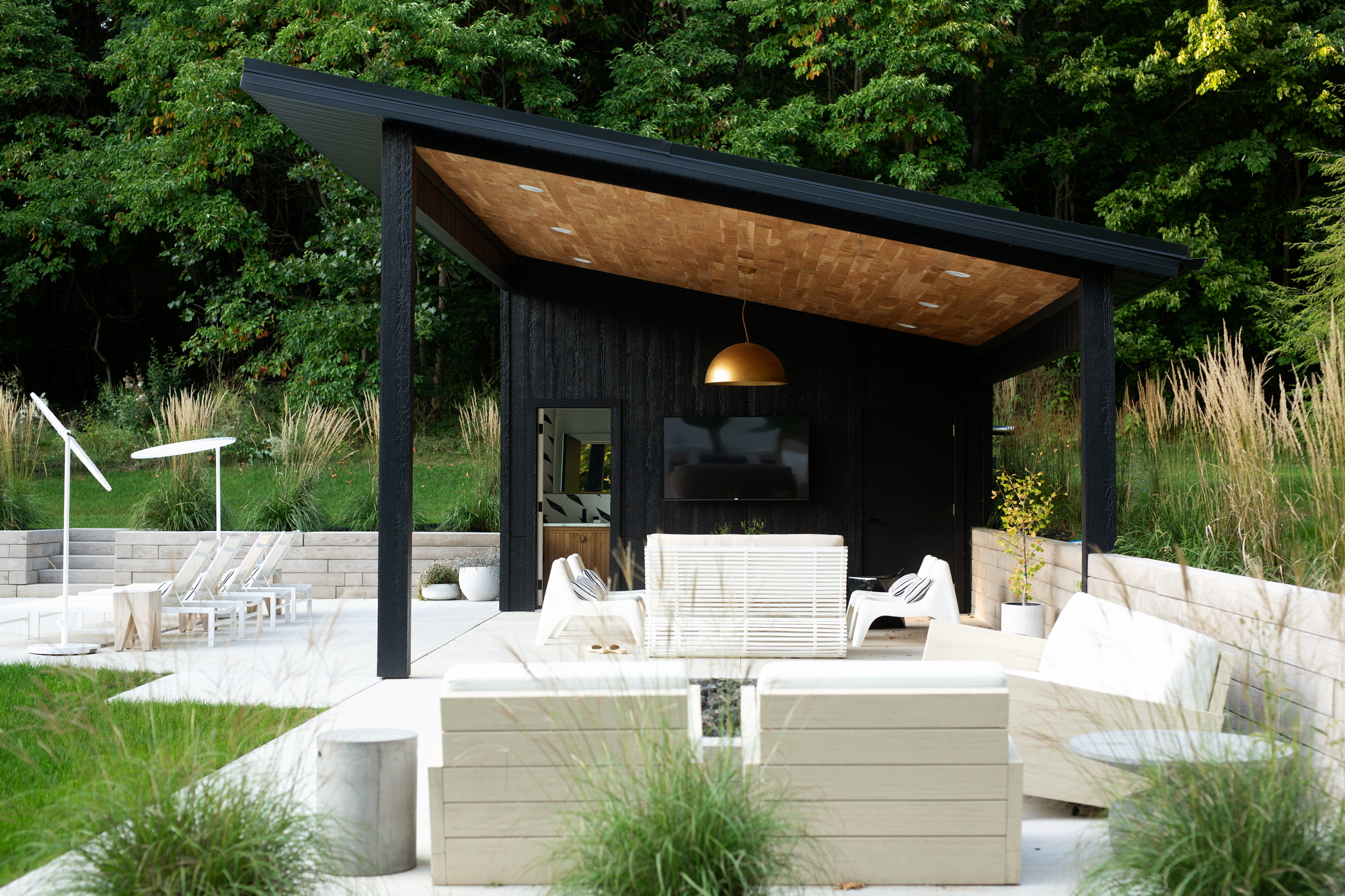 outdoor entertaining area under a slanted pergola with timber ceiling and pendant light with white outdoor chairs in three different seating arrangements and shrubs