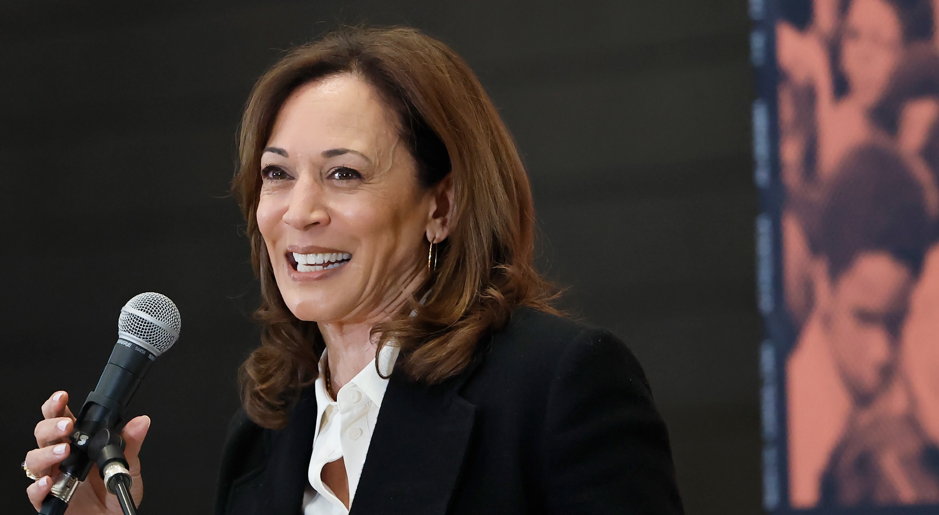 Kamala Harris in a white shirt and black blazer, giving a speech at a podium