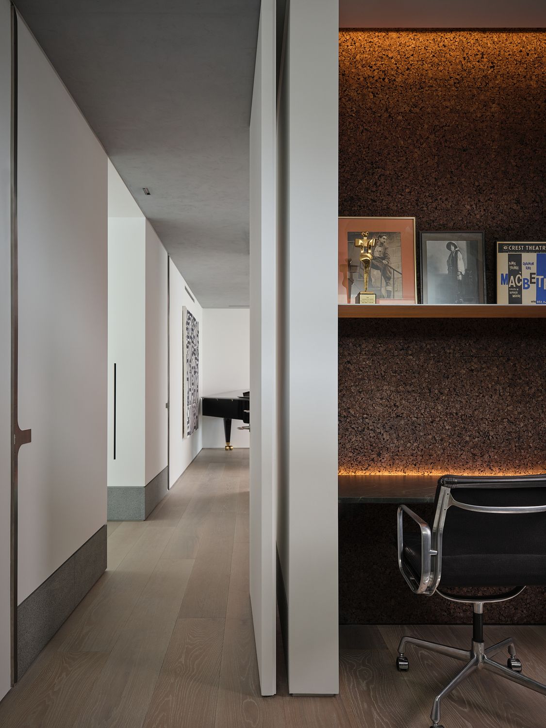 home office with cork wall and floating shelves with light timber floorboards next to a hallway looking down to a piano
