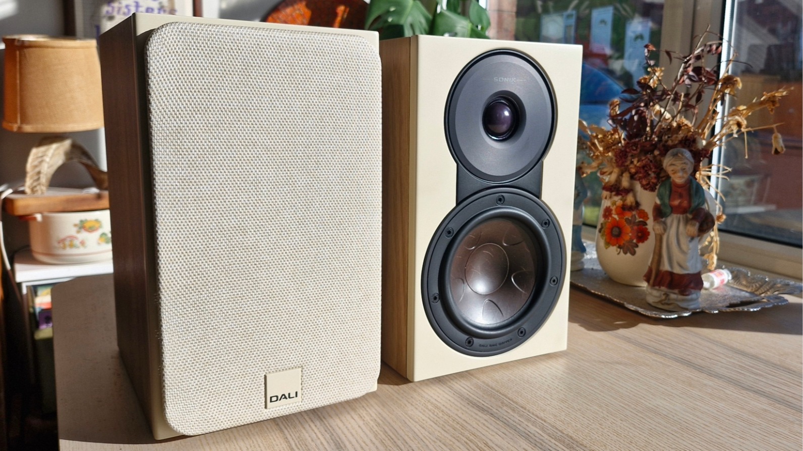 Dali Sonik 1 bookshelf speakers, one with and one without the detachable grille, on a wooden surface.