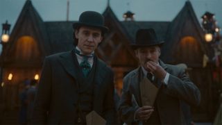 Image from the Marvel T.V. show Loki, season 2 episode 3. Two smartly dressed gentlemen each wearing a black hat and looking directly at the camera. The man on the right is eating a snack. Behind them is a large wooden building.