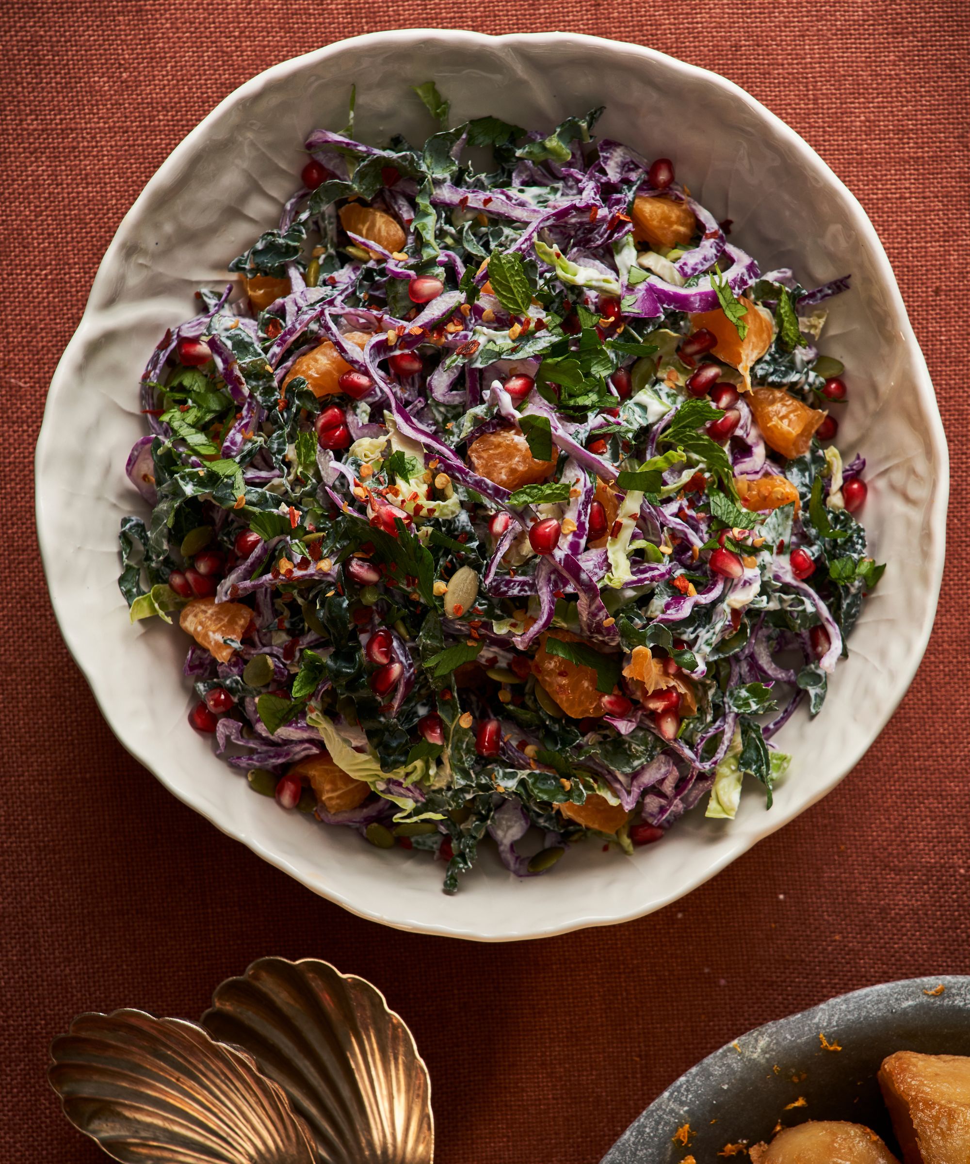 Bowl of winter slaw with red cabbage, oranges and pomegranate on a red tablecloth with gold salad servers
