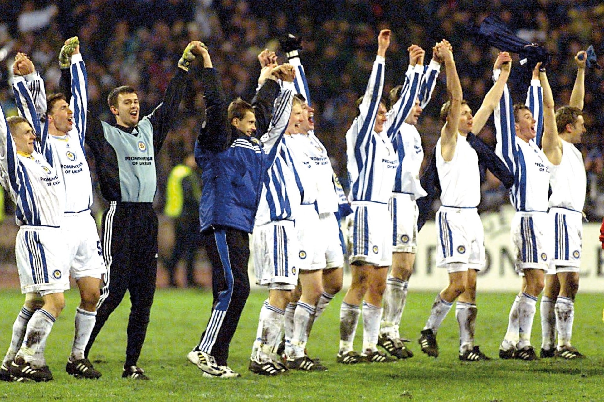 A group of football players in white and blue kits celebrating with their arms raised, showcasing their joy after a significant victory.