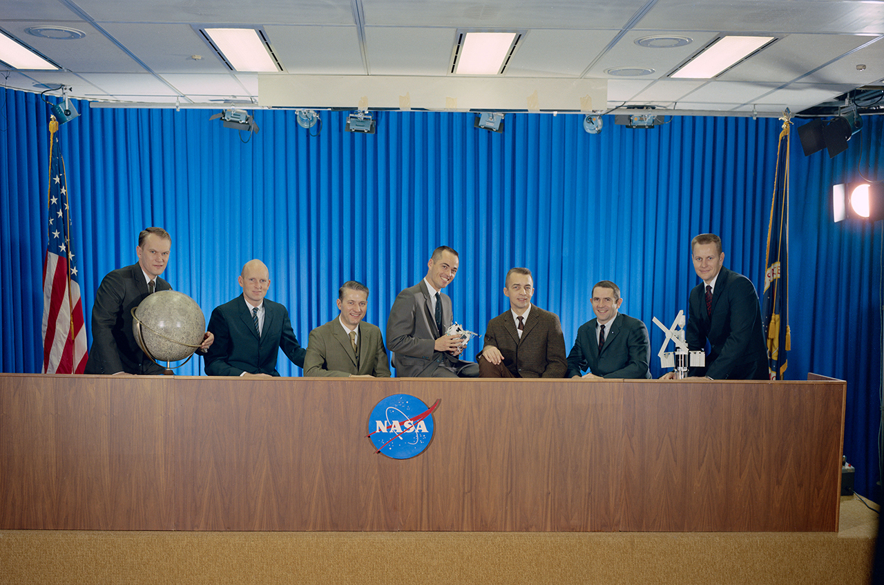 seven men in suits sit at a table with the nasa logo.