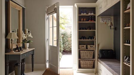 A bright entryway with an open glass-frame door, black side table with mirror above to the left, with lamps and flowers on it, and shelves and a bench to the right.