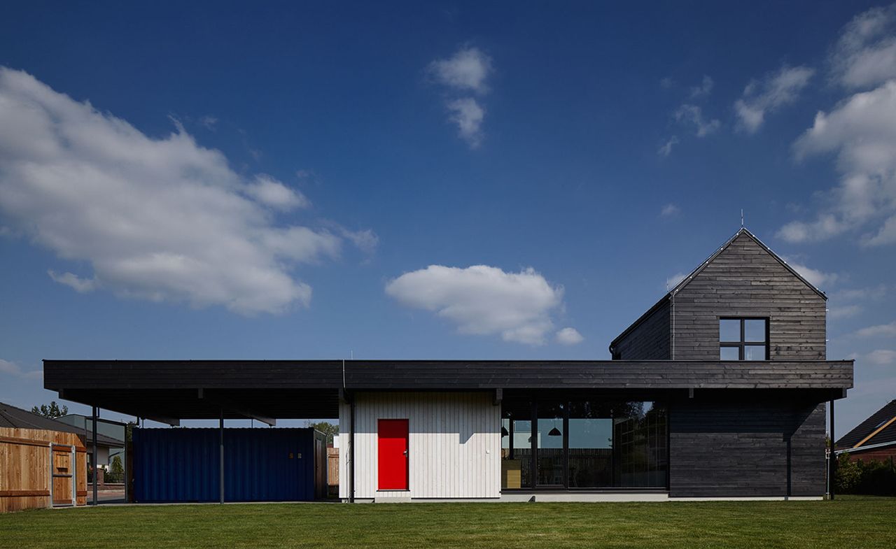 The exterior of a residential home clad in black timber planks with tall pitched-roof unit at one end of the structure surrounded by grey concrete and green lawn. The middle of the structure decorated in white wood with a red door and a blue container on the left shielded by the timber plank roof