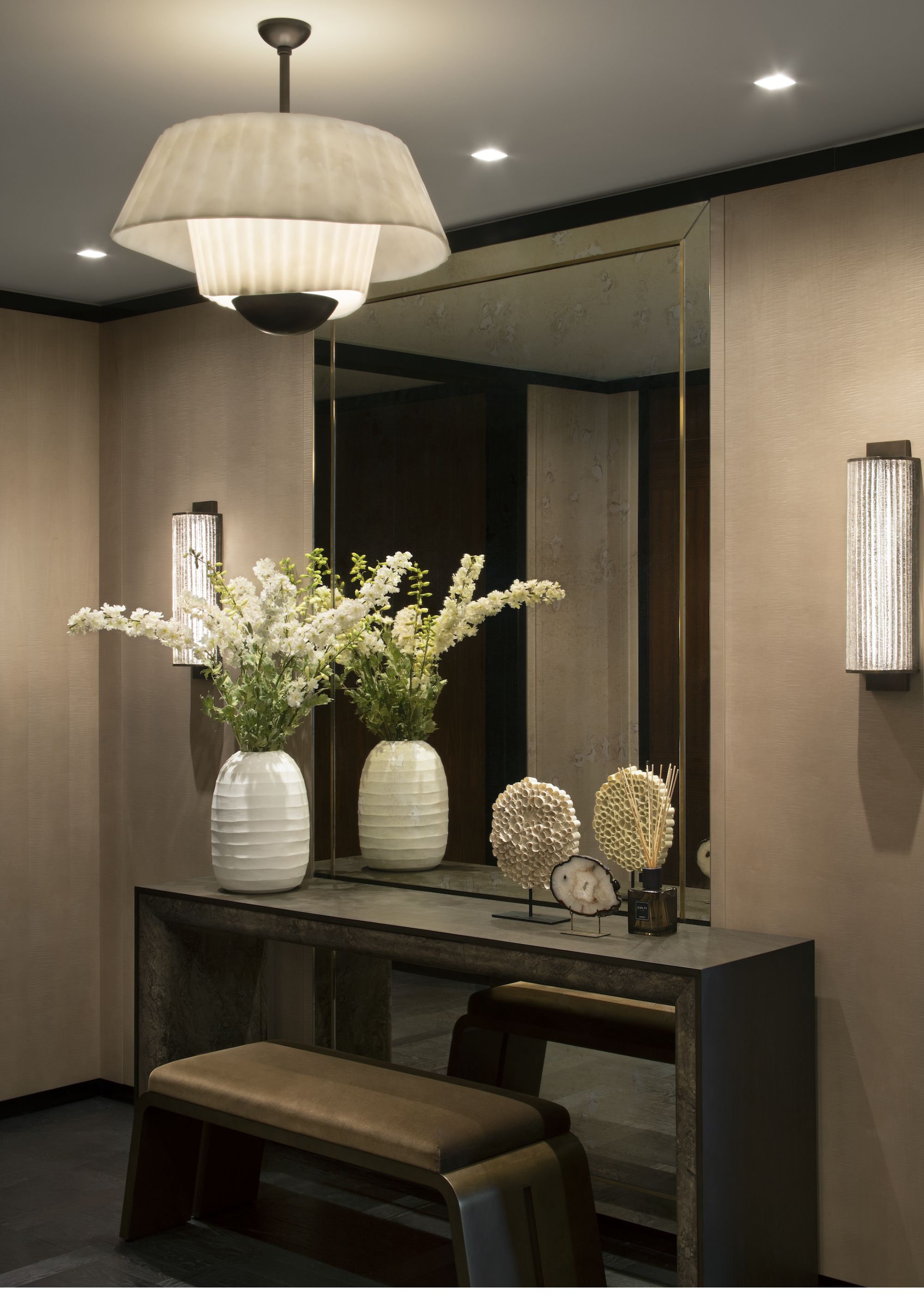 A table with a bench beside it and a large mirror above it. There is also a large ceiling lighting hanging above the table as well as a view of a wall lights on either sides of the mirror