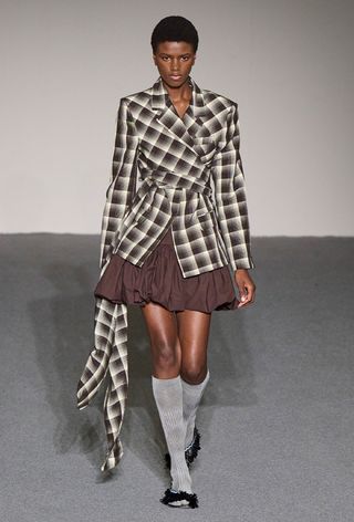model wearing stylish outfit with socks, including a brown and white plaid blazer with a brown ballon skirt and gray socks and sandals