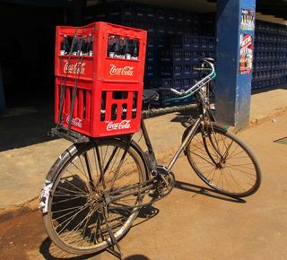 A bicycle packed outside with two filled red coca cola crates attached (behind the seat). photographed outside a building during the day