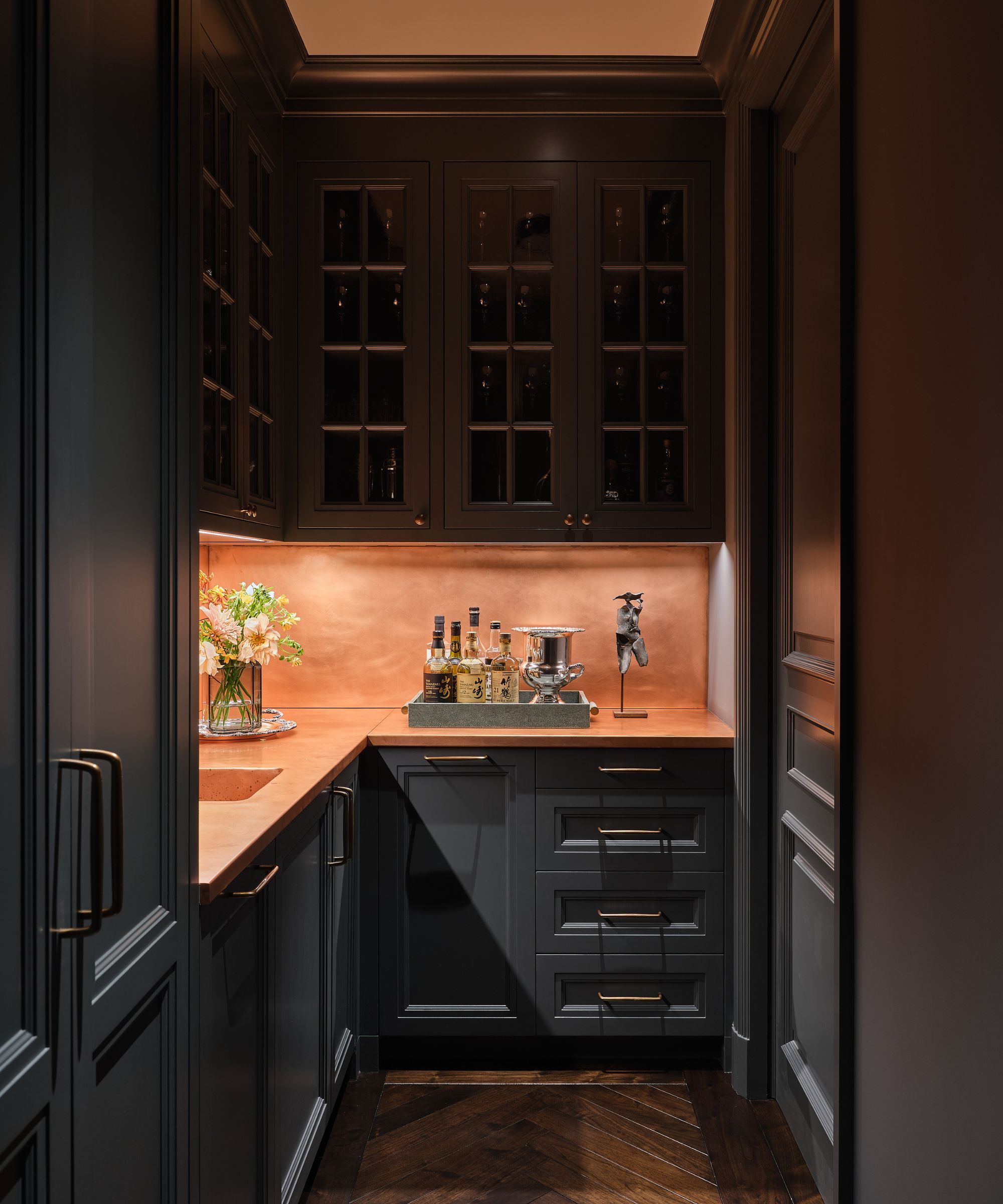 A scullery with dark gray cabinets, copper countertops and fittings, with under-cabinet lighting.