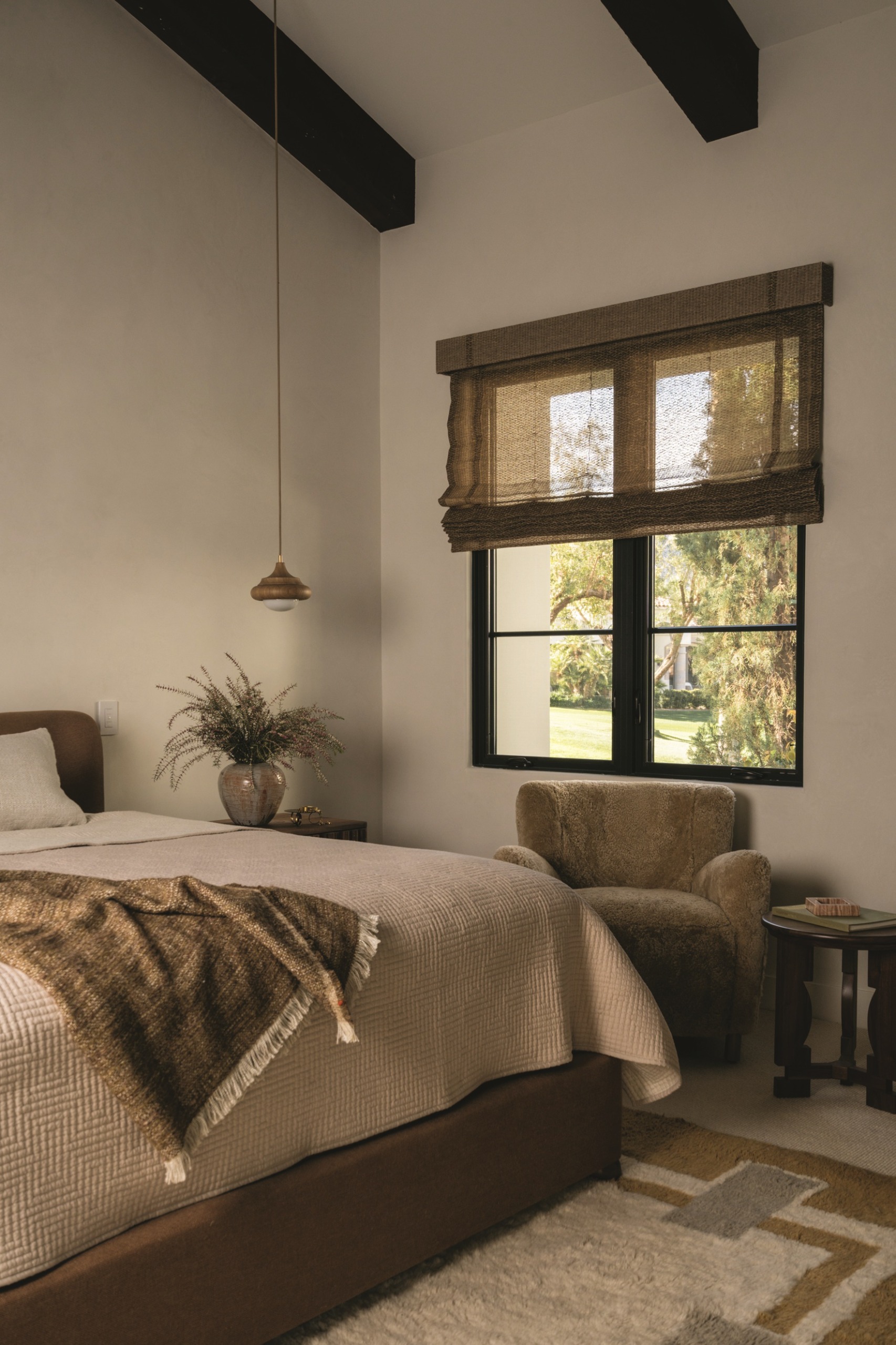 neutral bedroom with sheer blinds, shearling armchair, bed with waffle knit, exposed ceiling beams and hanging pendant light