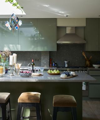 Modern kitchen with skylight, green cupboards and kitchen island, stone countertops, upholstered bar stools, hanging decorative mobile from ceiling