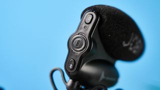 a black shotgun microphone by joby called the wavo pro is photographed by itself against a blue background, showing its muff, dead cat, attachment foot, and connection ports