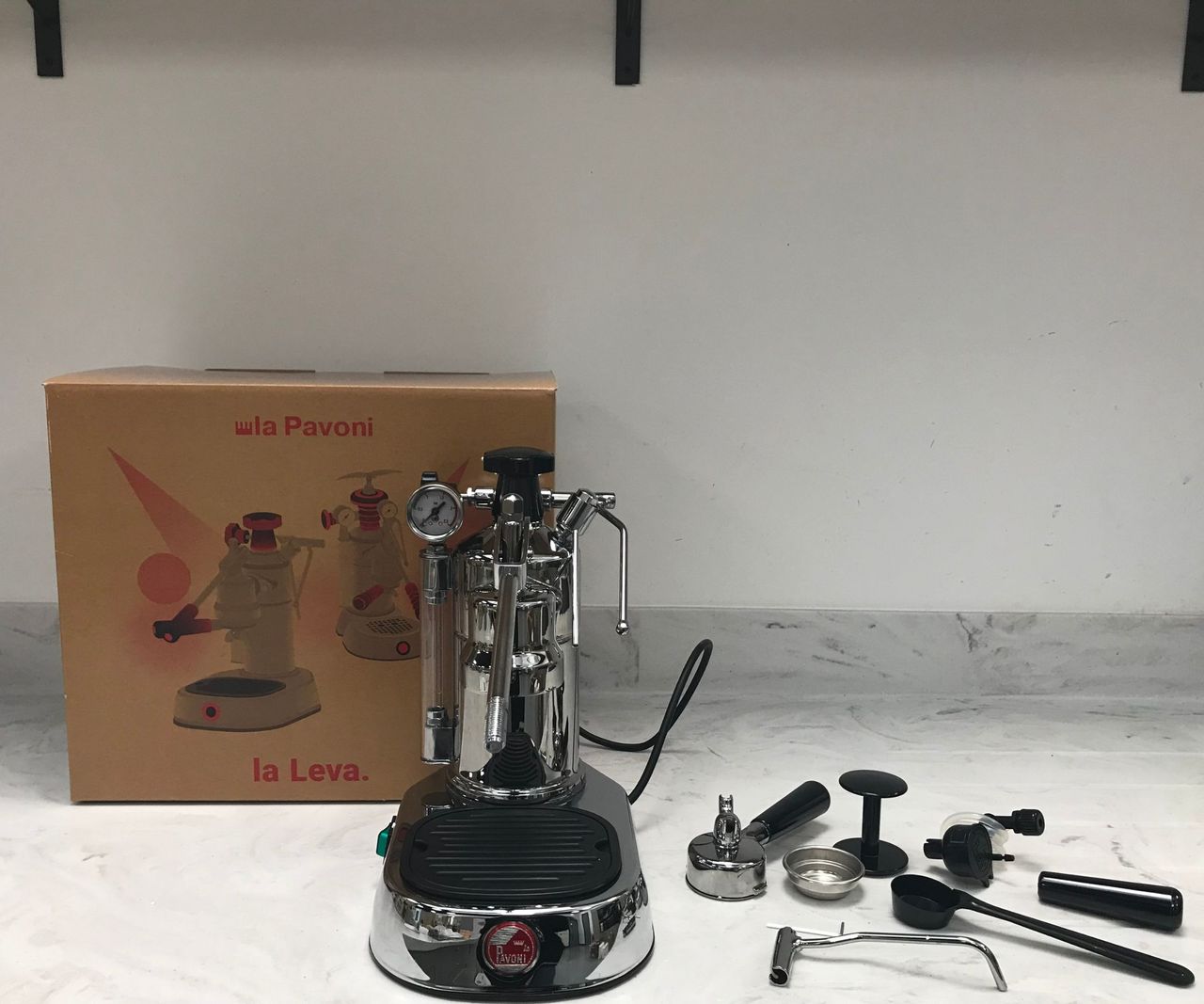 La Pavoni professional unboxed on countertop with all the small components needed to make espresso. The brown box sits behind it on a white countertop.