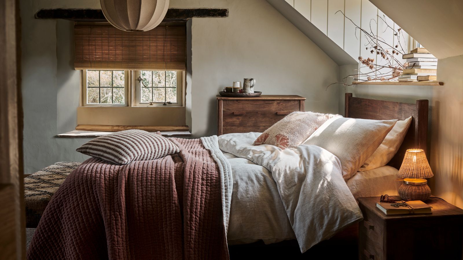 A bed in an attic room, with layered white and rust bedding. A warm toned wicker lamp n the table beside it. A large window partially covered by a rattan blind in the background. 