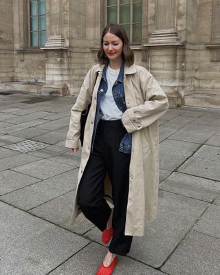 Fashion influencer Marissa Cox poses on the streets of Paris in a trench coat outfit with a white tee, denim jacket, black pants, and red mesh flats