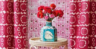 Close up shot of pink patterned curtains hanging in front of a pink patterned wallpaper with a blue patterned vase on a woven side table to show maximalist decor