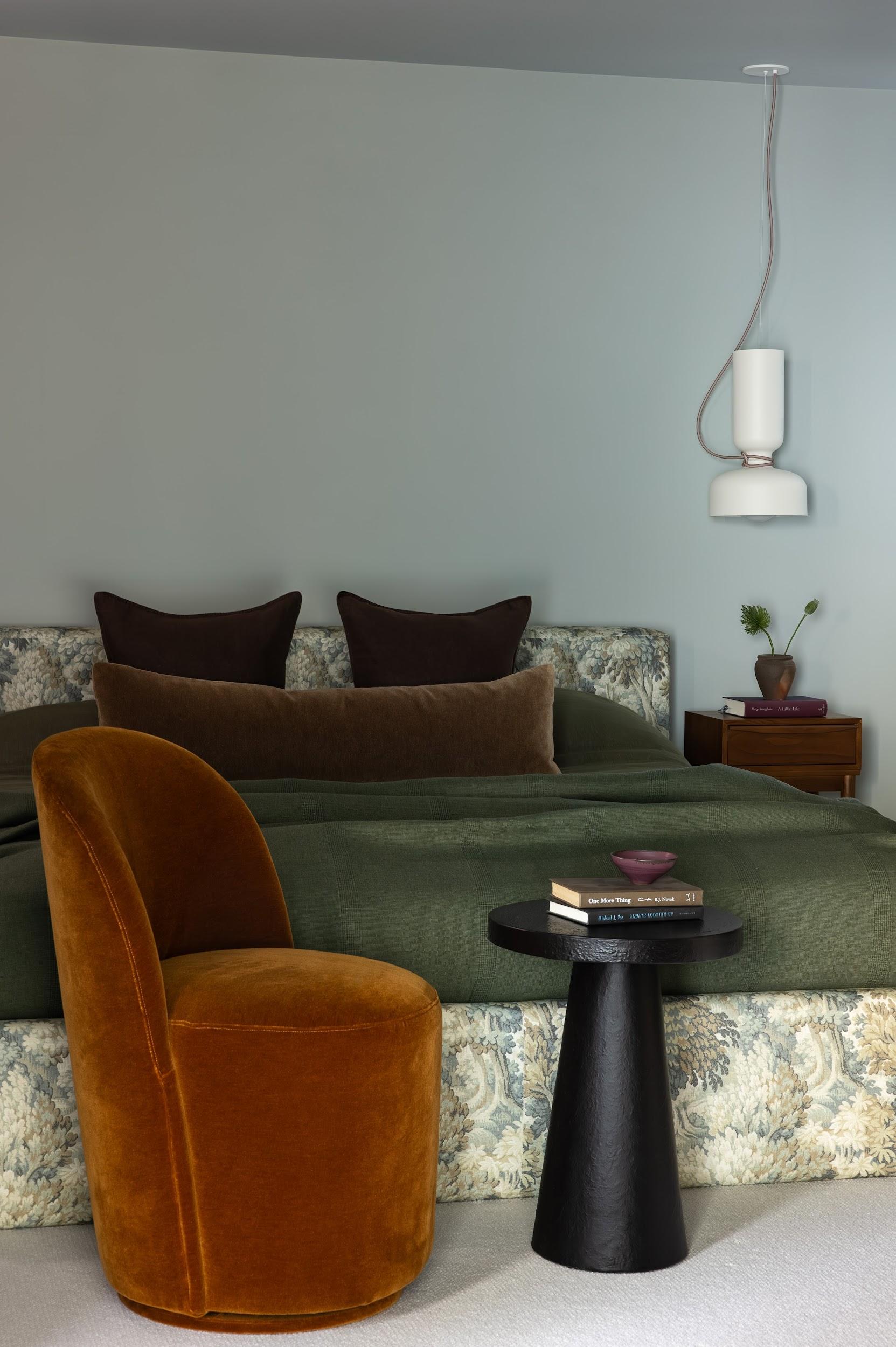 A modern bedroom with a floral bedframe, green bedding and a rust accent chair at the foot of the bed next to a round black side table; a white pendant light hangs from the ceiling above the bedside