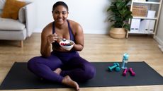 A woman sits cross-legged on a yoga mat with small dumbbells and a water bottle next to her. In her hands is a bowl of cereal and fruit.