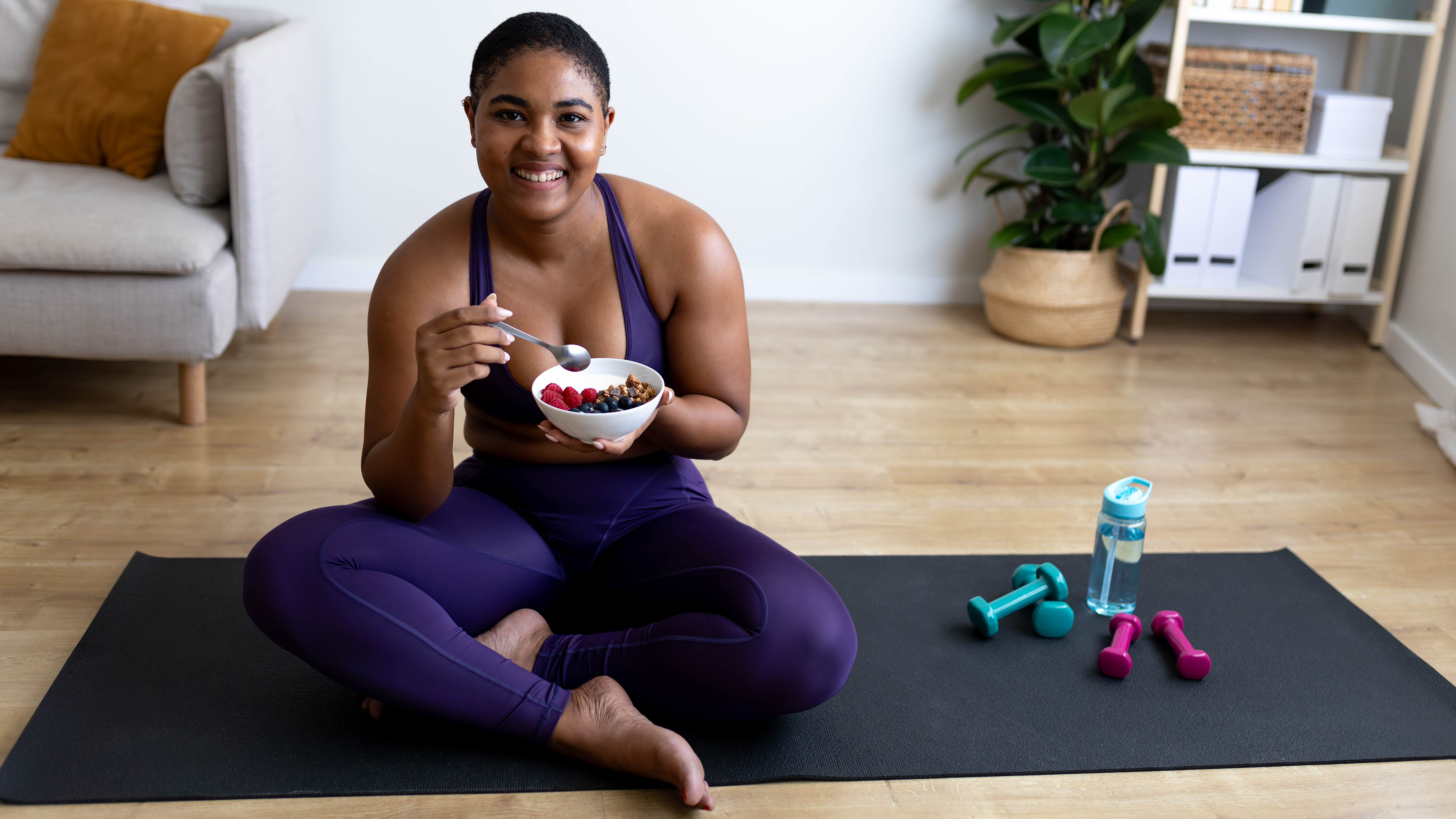 A woman sits cross-legged on a yoga mat with small dumbbells and a water bottle next to her. In her hands is a bowl of cereal and fruit.