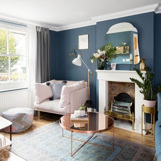 Living room with rug on wooden floor, blue walls, mirror over fireplace, round coffee table with mirror finish and upholstered armchair next to window